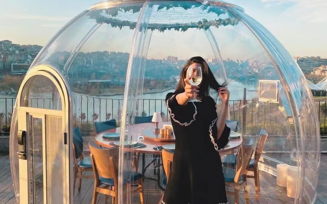 Woman taking a photo with her smartphone in front of the AstroSphere Retreat luxury stargazing dome with a cityscape in the background.