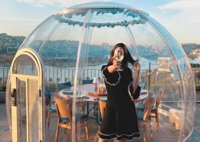 Woman taking a photo with her smartphone in front of the AstroSphere Retreat luxury stargazing dome with a cityscape in the background.