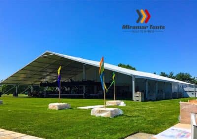 Large white All-Occasion Canopy Tent set up in a grassy area on a sunny day, with blue sky above and colorful flags at the entrance.