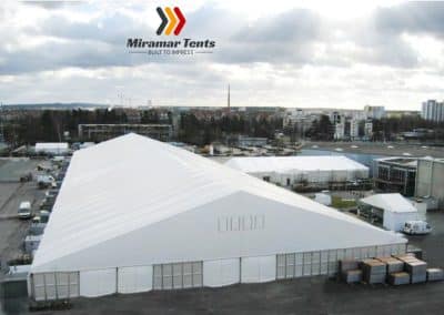 A large white All-Occasion canopy tent in an open outdoor area with urban buildings in the background under a cloudy sky.