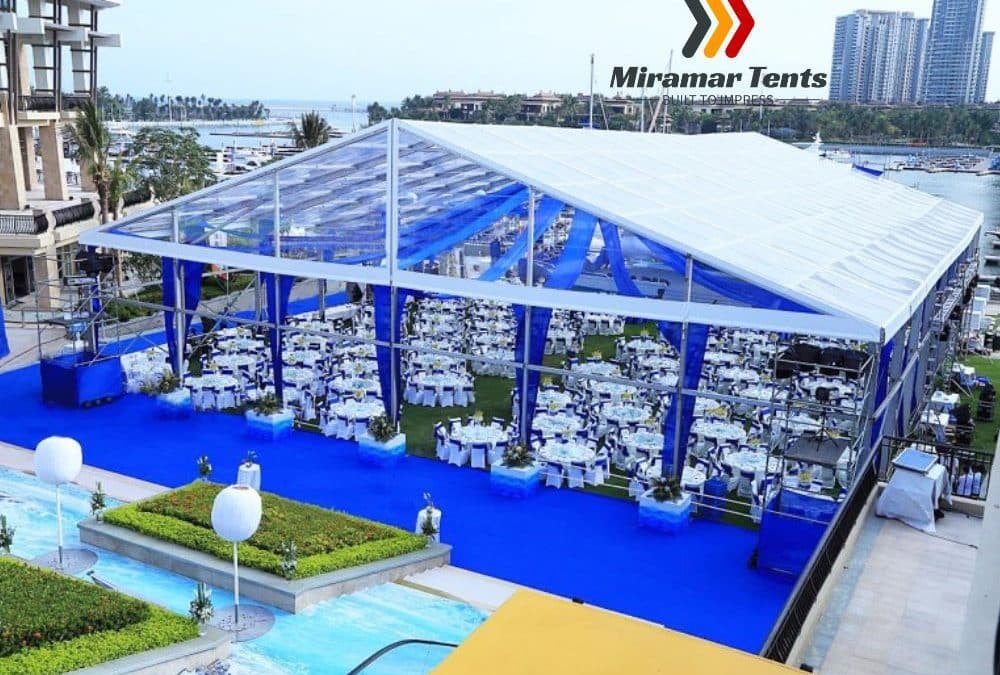 Sentence with replacement: Outdoor event setting with a large All-Occasion Canopy Tent, white chairs and tables, set on a blue carpet near a waterfront, with cityscape in the background.