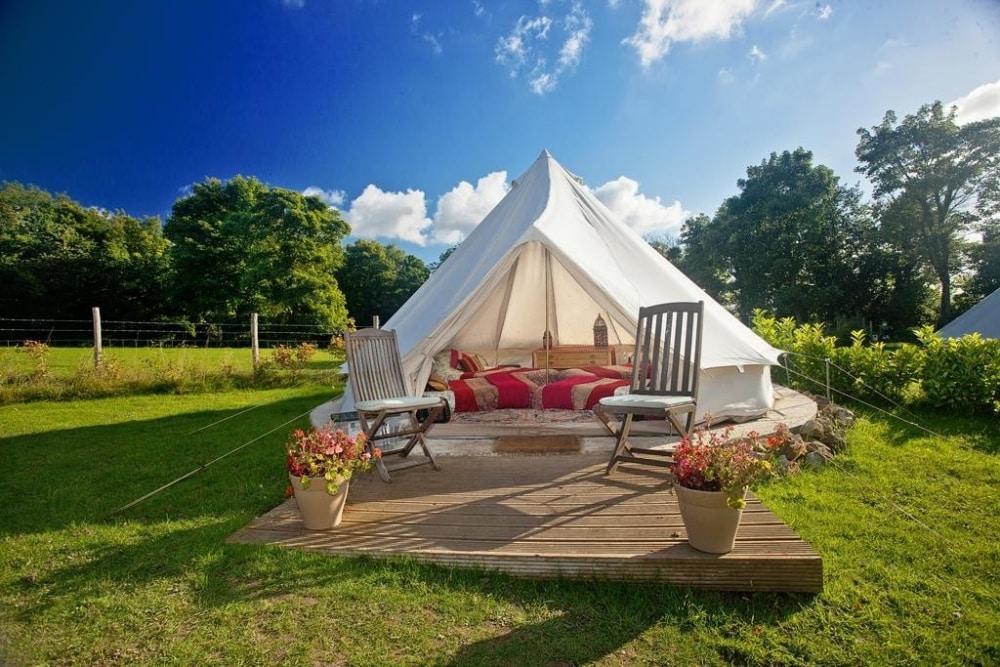 A luxurious glamping **Bell Tent** with comfortable seating and floral accents set in a sunny, grassy area with trees.