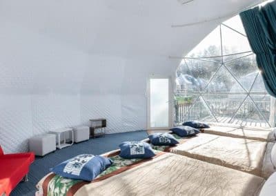 Interior of a white geodesic dome tent with three beds adorned with pillows and blankets, a red couch, two small tables, and a view of the outdoors through large windows.