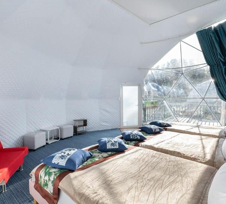 Interior of a white geodesic dome tent with three beds adorned with pillows and blankets, a red couch, two small tables, and a view of the outdoors through large windows.