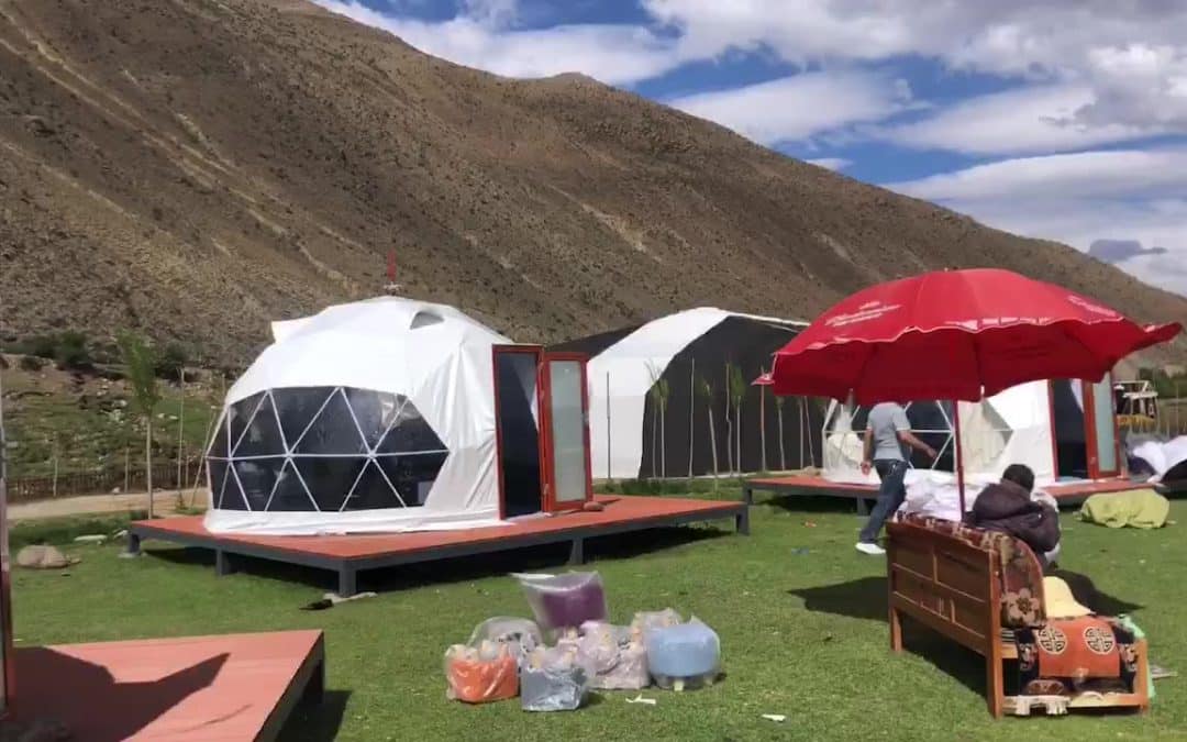 Outdoor scene with white geodesic dome tents on wooden platforms, set against a mountainous backdrop. Some outdoor furniture and a red umbrella are visible, with people and scattered items around.