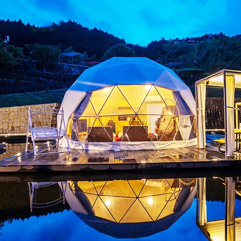 A lit dome-shaped glamping tent is reflected in a body of water at dusk, surrounded by a wooden deck and a swing. Trees and hills are visible in the background under a clear sky.