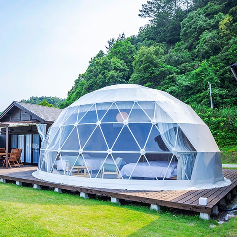 A transparent geodesic dome with furnishings inside is situated on a wooden deck, surrounded by greenery and adjacent to a traditional wooden house.