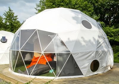 A white geodesic dome tent sits on a wooden platform surrounded by greenery. It has circular windows and a large, transparent entry with a visible sleeping bag and pillows inside.