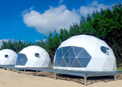 Three white geodesic dome tents set on a sandy beach with green trees in the background and a blue sky above.