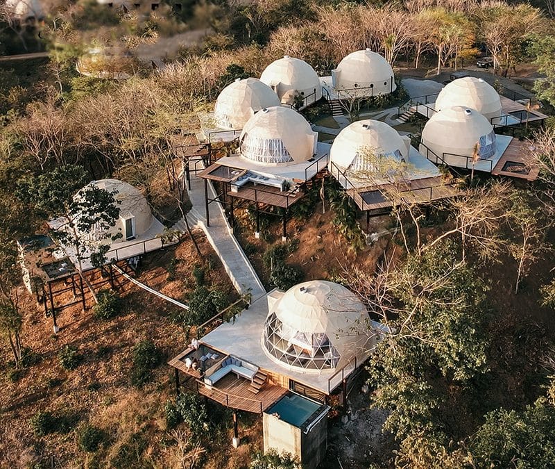 Aerial view of a complex with multiple dome-shaped buildings, resembling glamping dome tents, surrounded by trees. Wooden pathways connect the structures, some of which feature small pools.