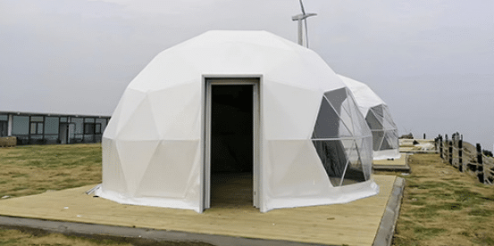Two Glamping Dome Tents with geometric designs stand on wooden platforms in a grassy area, accompanied by a wind turbine in the background. Perfect for glamping enthusiasts, these structures blend modern aesthetics with nature.