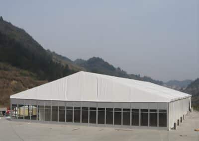 A large white marquee tent with transparent lower walls set up on a concrete surface, with mountains and foggy sky in the background.