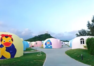 Colorful, dome-shaped buildings with cartoon character designs, surrounded by greenery and a paved walkway, with a hill and tower in the background under a clear sky.