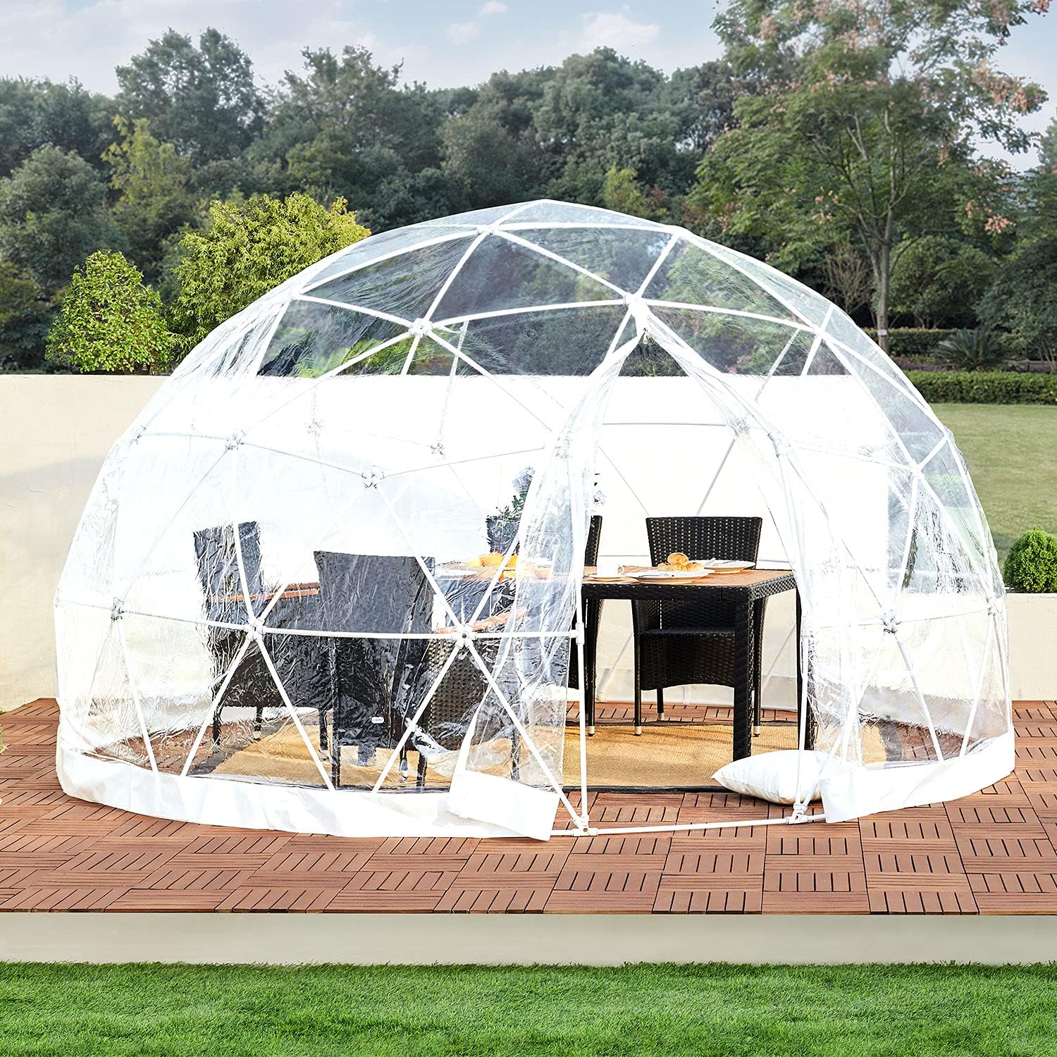 A clear geodesic dome structure encloses a patio dining set with black chairs and a table on a wooden deck, situated in an outdoor garden area.