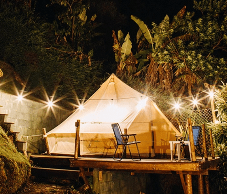 A well-lit tent on a wooden platform is surrounded by string lights and plants at night. Two chairs and a small table are placed in front.