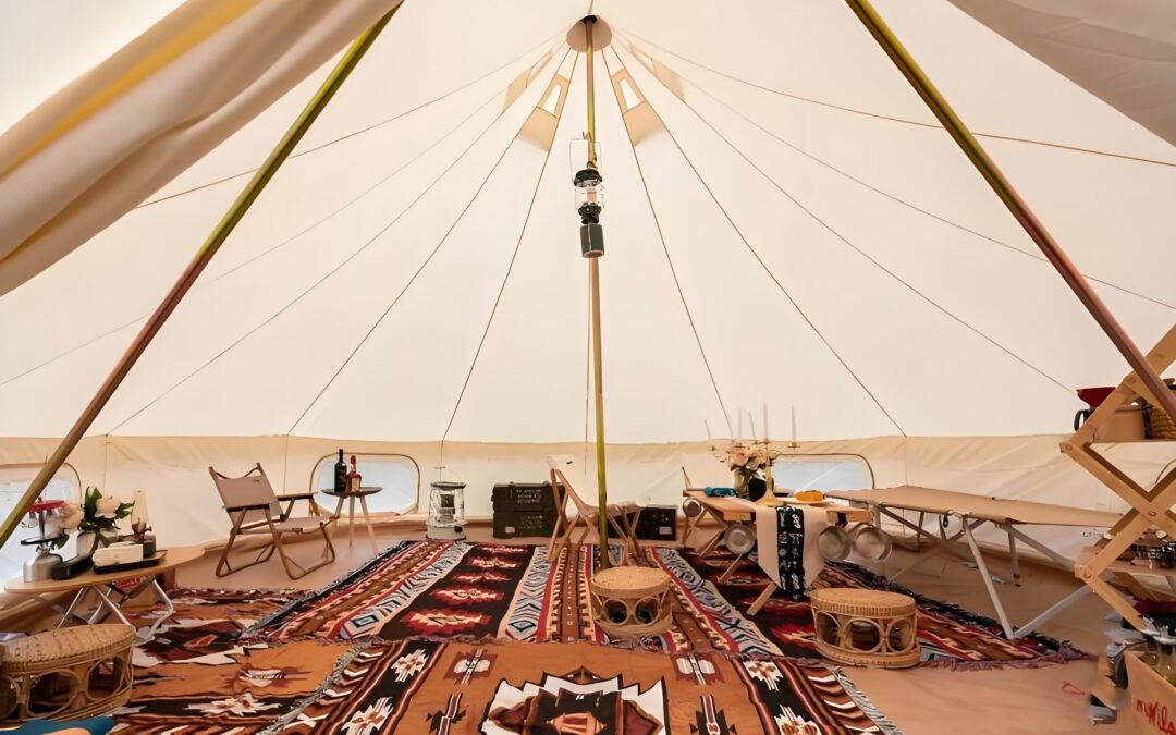 Interior of a spacious tent with colorful rugs, chairs, tables, and decorative items, including a hanging lantern in the center.