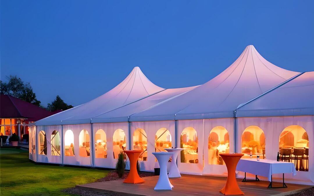 A large white party tent illuminated from within sits gracefully on the grass, with tables outside adorned in vibrant orange and white cloths.