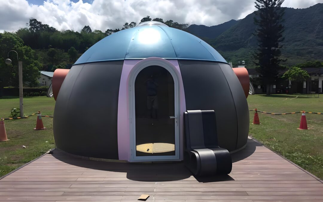 A small, dome-shaped structure with a blue and gray exterior, featuring a glass door and a black exterior air conditioning unit; situated on a wooden platform, with mountains and trees in the background.