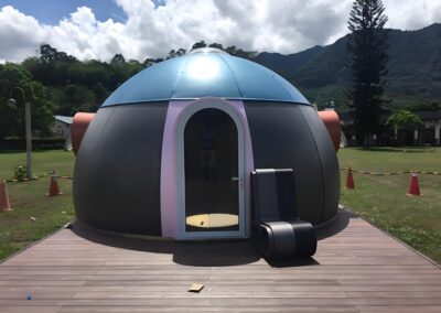 A small, dome-shaped structure with a blue and gray exterior, featuring a glass door and a black exterior air conditioning unit; situated on a wooden platform, with mountains and trees in the background.