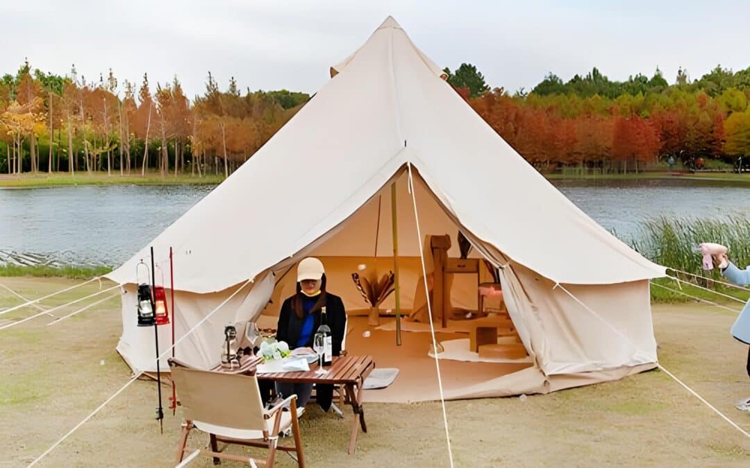 A person sits at a table with camping gear outside a large beige tent by a lake, surrounded by autumn trees.