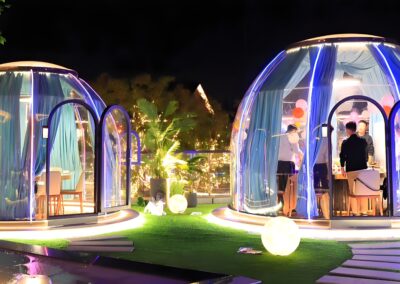 Two illuminated glass igloo-style dining pods with people inside at night, decorated with fairy lights and balloons.