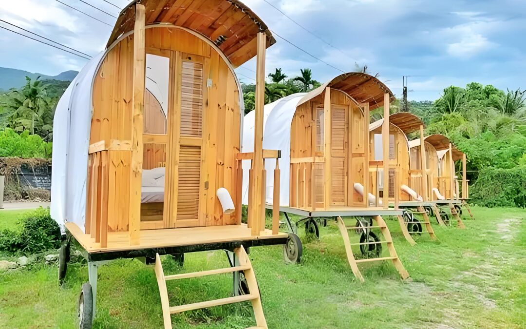 Four wooden caravan-style tiny houses on wheels with curved roofs, resembling a quaint carriage tent, are lined up on grassy ground. Steps lead to each inviting entrance.