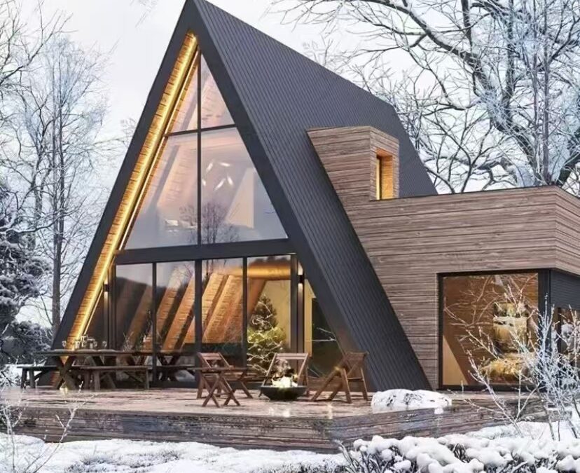 A-frame cabin with large windows, wooden exterior, and snowy surroundings. Outdoor seating is on a wooden deck, and trees are visible in the background.