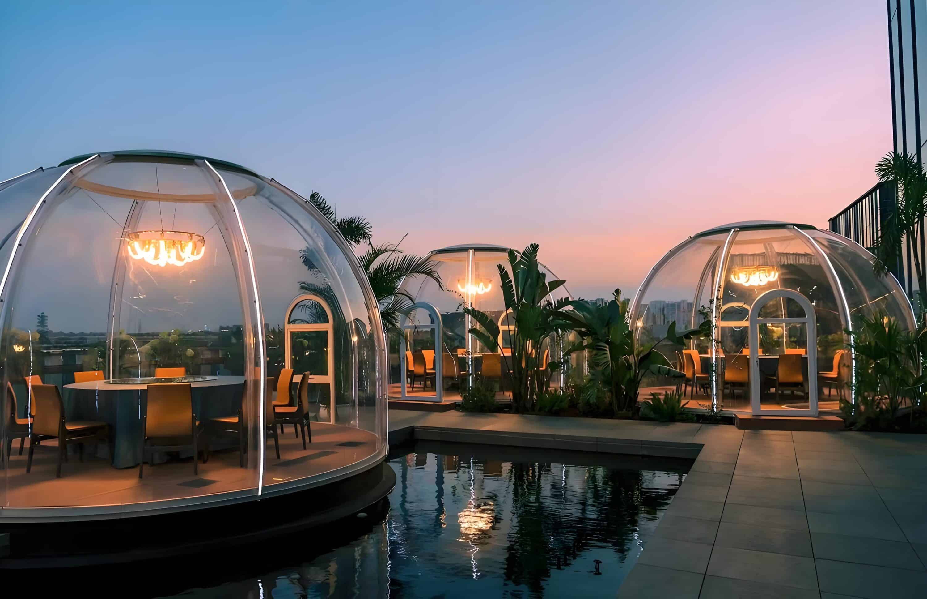 Outdoor dining area with transparent igloo-like domes, each containing a set dining table and chairs, beside a water feature at sunset.