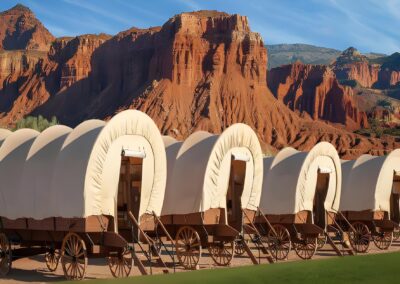 A row of covered wagons is set against a backdrop of red rock cliffs and blue sky.