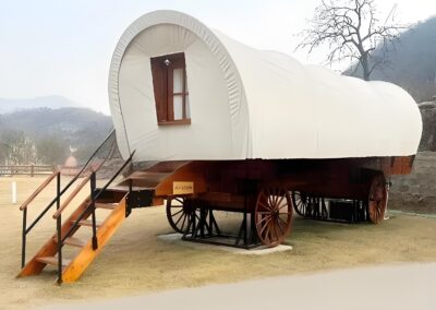 A white covered wagon on large wooden wheels with a set of wooden steps leading to the entrance stands on a lawn with hills and a bare tree in the background.