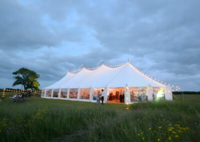 A large white event tent with string lights is set up in a grassy field under a cloudy sky. People are visible inside the tent.