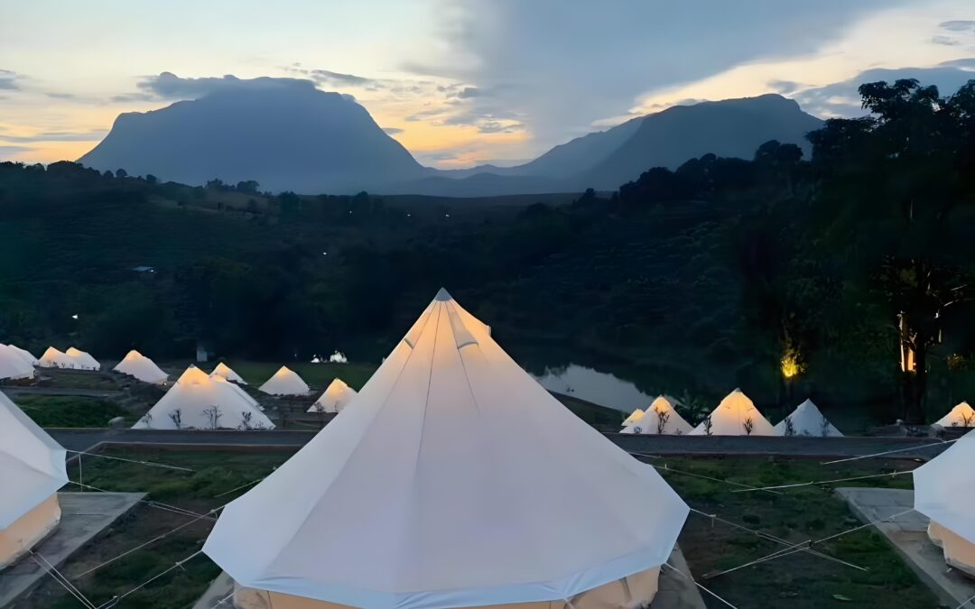 A row of illuminated bell tents in a campsite at dusk, set against a backdrop of mountains and a partly cloudy sky.