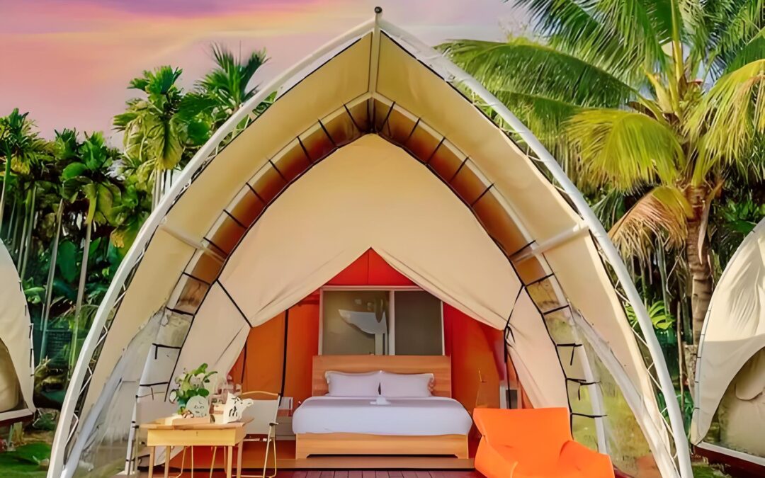 A sailboat tent sits on a raised deck, offering a luxurious glamping experience with a bed, table, and orange chair. Surrounded by palm trees under the vibrant hues of a sunset sky.