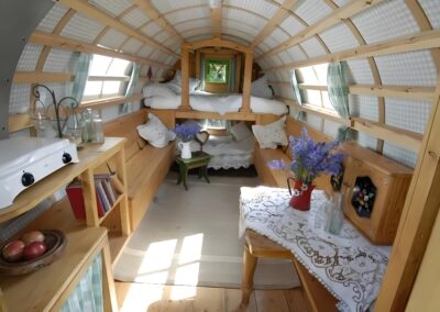 Interior of a cozy, wood-paneled camper with a small kitchen, table, bench seating, and a bunk bed. It's decorated with flowers and lace accents, creating a warm, homey atmosphere.