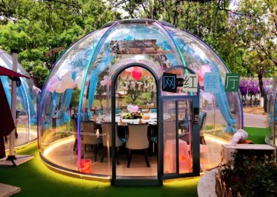 A transparent dome-shaped dining area with blue accents and string lights, set up outdoors under green trees. The interior includes a dining table and chairs, with some decorations present.