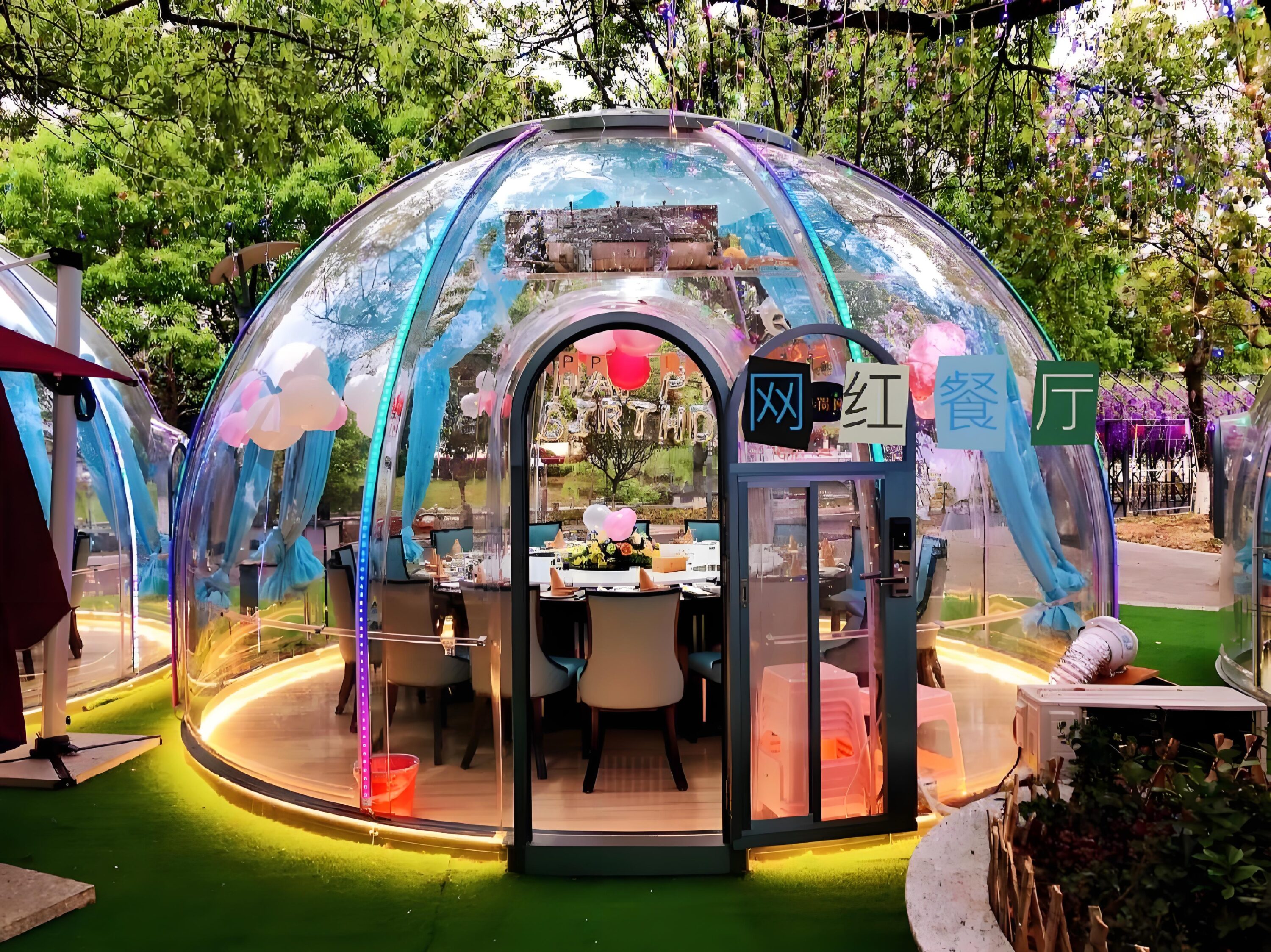 A transparent dome-shaped dining area with blue accents and string lights, set up outdoors under green trees. The interior includes a dining table and chairs, with some decorations present.