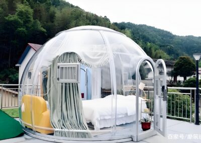 A transparent dome-shaped room with a bed, armchair, and curtains is situated on a wooden deck in a mountainous area.