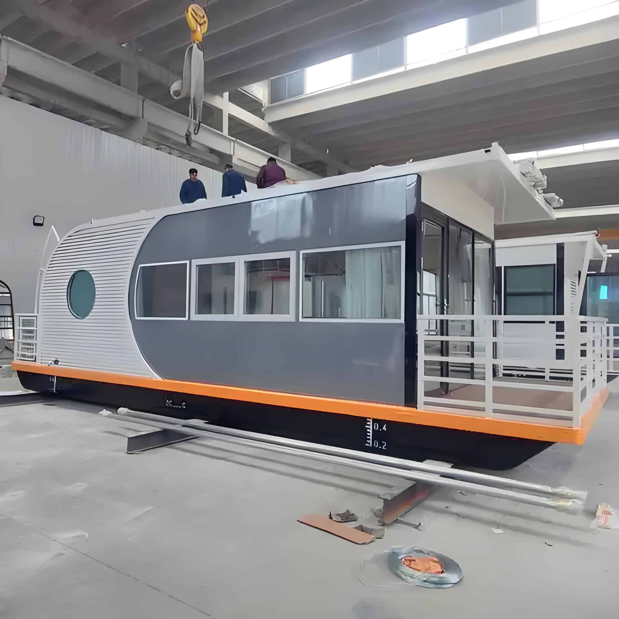 A modern houseboat in a warehouse under construction, featuring large windows and a white and gray exterior with orange trim. Some people are visible on the roof, possibly working or inspecting.