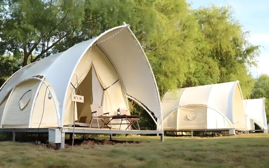 Two white canvas tents on wooden platforms in a grassy area surrounded by trees, with outdoor seating in front of one tent.