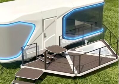 Futuristic white travel trailer with blue LED accents, featuring large windows and a small wooden deck attached to the side equipped with steps and railings, situated on a grassy area.