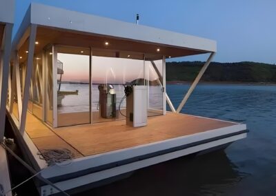 A modern floating structure with large glass windows and a wooden deck is positioned on a calm body of water at dusk. Hills are visible in the background under a clear sky.