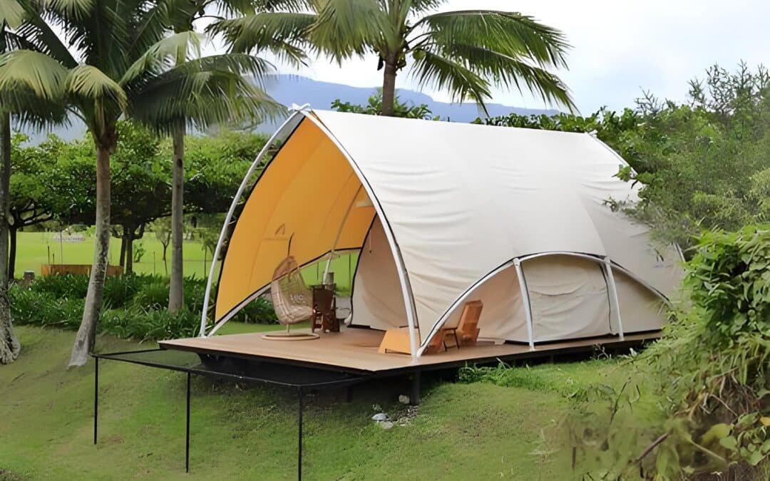 A glamping tent with a curved roof set on a wooden deck, surrounded by palm trees and greenery. It features a hanging chair and a lounge chair inside.