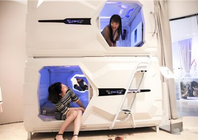 Two Women Are Inside Futuristic, White Sleeping Pods, With One Seated Inside The Lower Pod And The Other Looking Out From The Upper Pod.