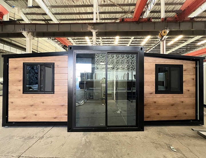 A Prefabricated Modular Building With A Central Glass Sliding Door And Two Side Windows, Situated Inside A Large Industrial Warehouse.