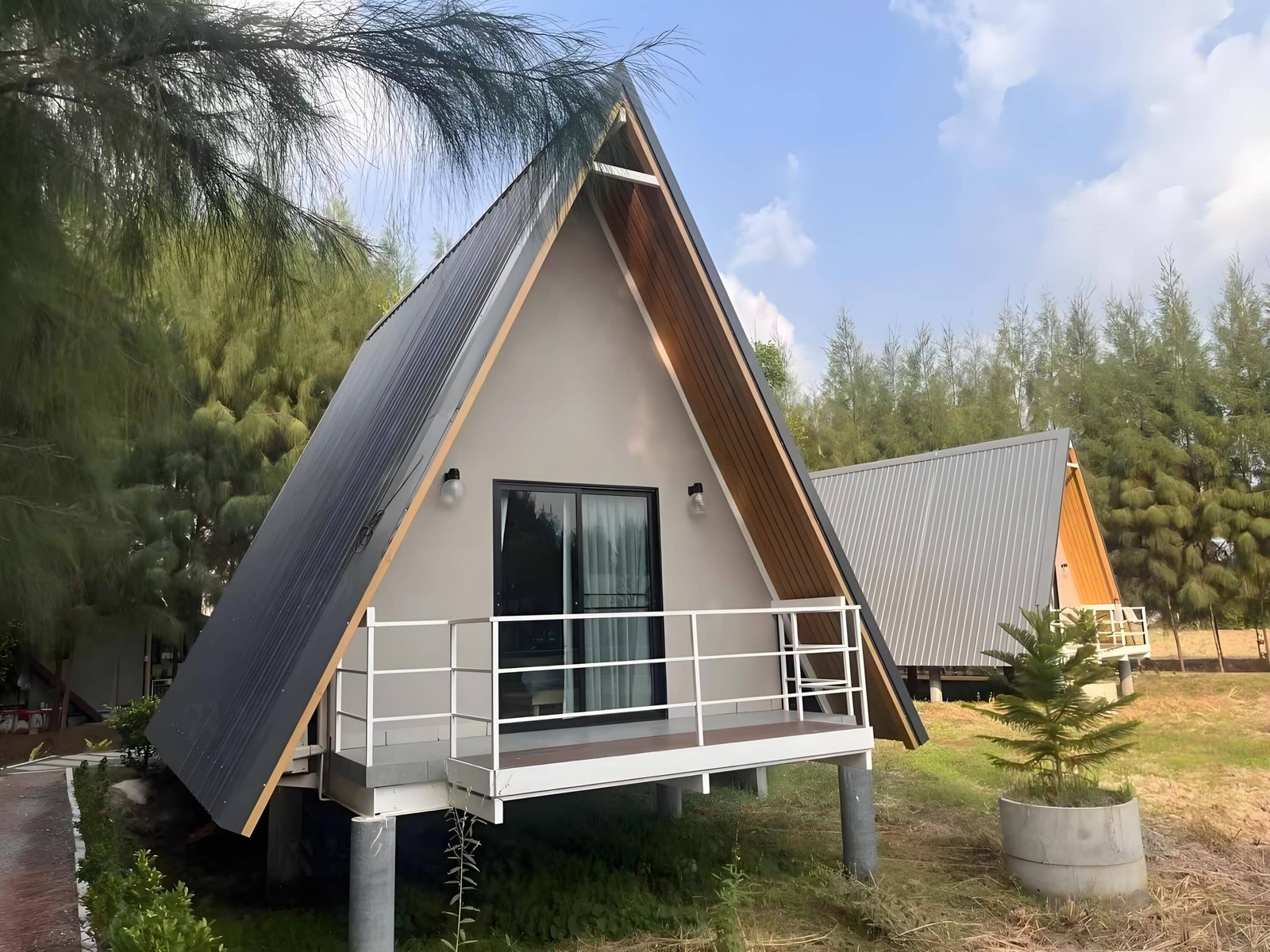 A Frame Cabin With A Metal Roof And A Small Porch, Surrounded By Trees And Sky In The Background.