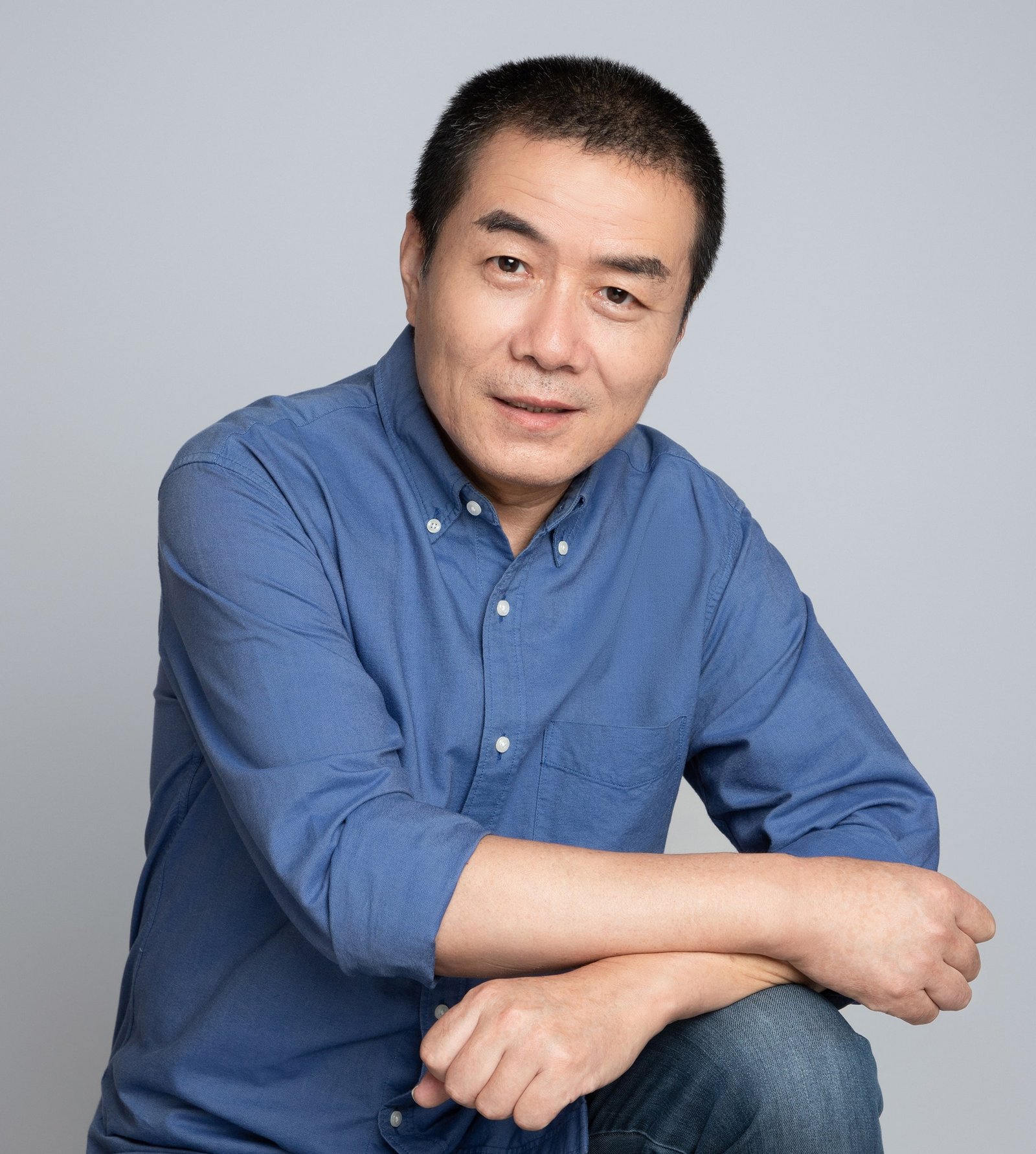 A Man In A Blue Button Up Shirt And Jeans Poses Against A Plain Gray Background.