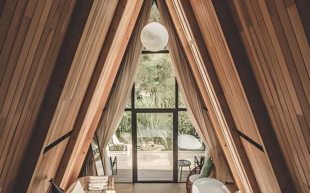 A Frame Cabin Interior With Wood Paneling. Features A White Sofa, Round Coffee Table, Clawfoot Bathtub, Wicker Chair, And Large Window Overlooking Lush Greenery.