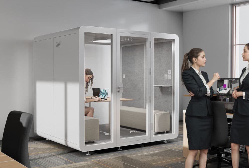 Two women in business attire talk near a glass walled office pod, where another person sits at a desk with a laptop in a modern open office environment.