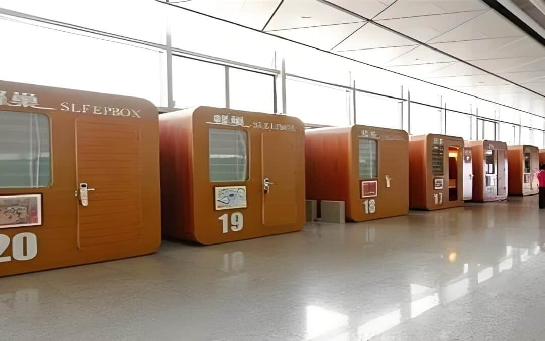 Row of numbered capsule beds with closed doors and frosted windows in a bright, modern indoor space; designed for traveler well being, one person stands nearby.