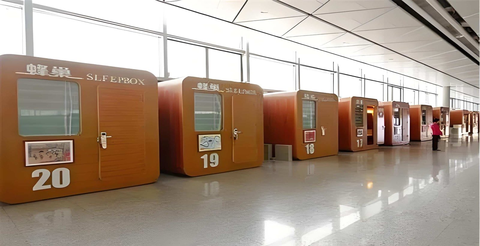 Row of numbered capsule beds with closed doors and frosted windows in a bright, modern indoor space; designed for traveler well being, one person stands nearby.